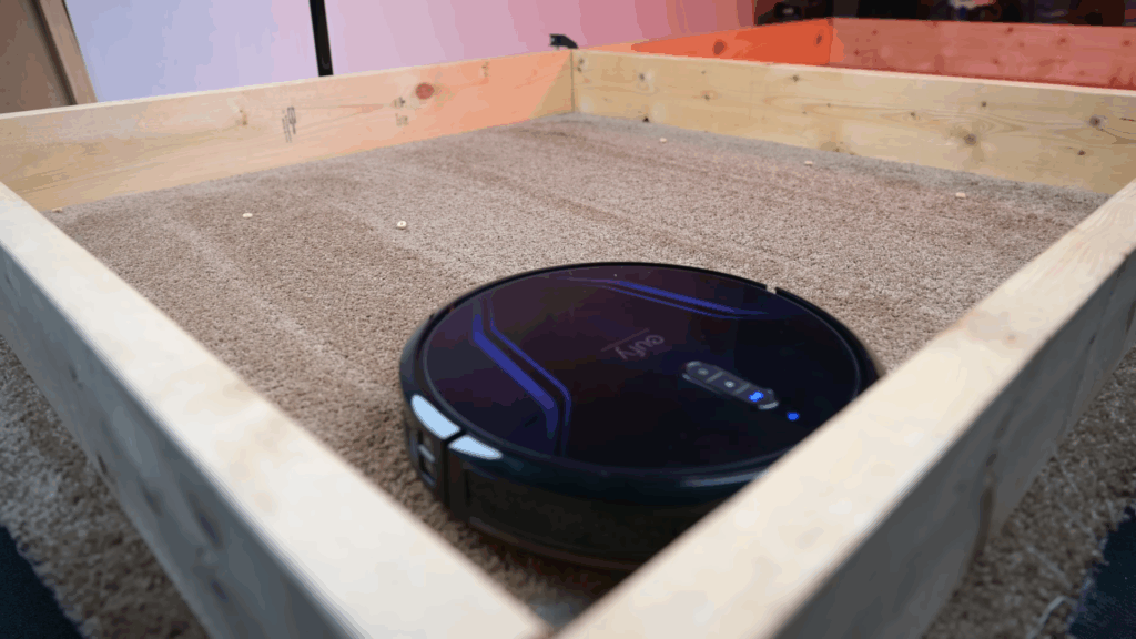 Eufy RoboVac G40+ navigating on a carpet surface during a debris pickup test inside a wooden test box.
