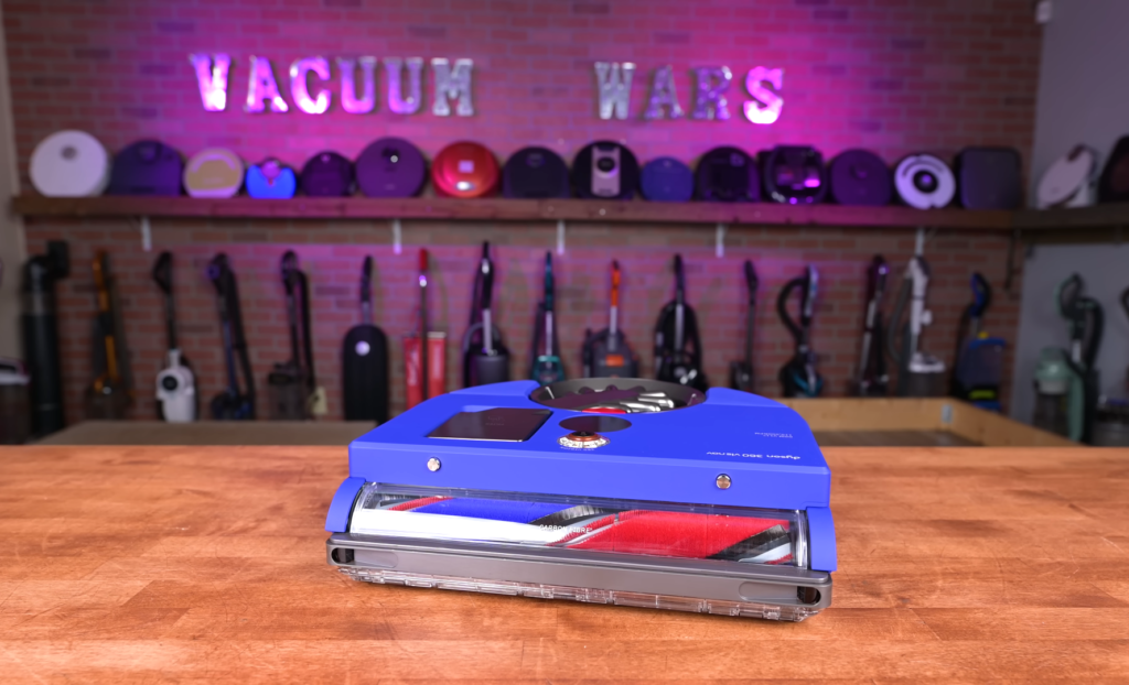 The Dyson 360 Vis Nav robot vacuum placed on a wooden surface with the Vacuum Wars logo illuminated in neon in the background.