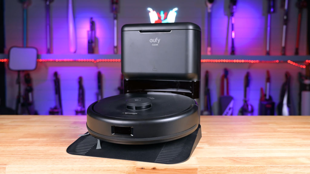 Eufy L60 robot vacuum on a wooden table, against a blurred background of vacuum cleaners on display and colorful lighting.