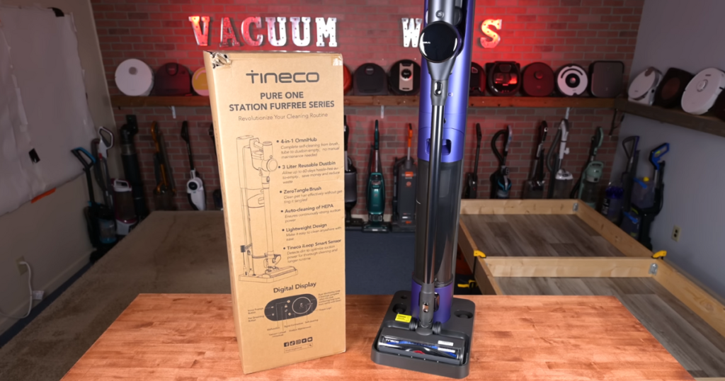 Tineco Pure ONE Station FurFree vacuum with its packaging box highlighting key features next to a collection of various vacuum cleaners in the background.