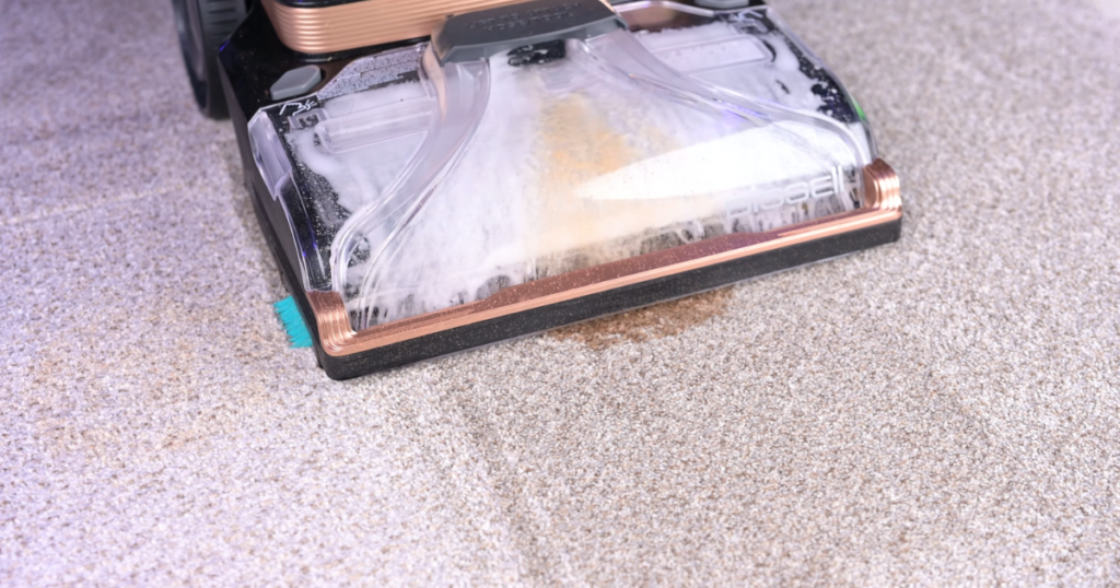 A carpet cleaner in action, with the cleaning head moving over a stained area of carpet, visibly removing the stain and cleaning the carpet fibers.