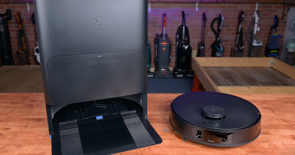 Ecovacs Deebot T30S robot vacuum placed next to its Omni Station on a wooden surface, with various vacuum cleaners in the background.