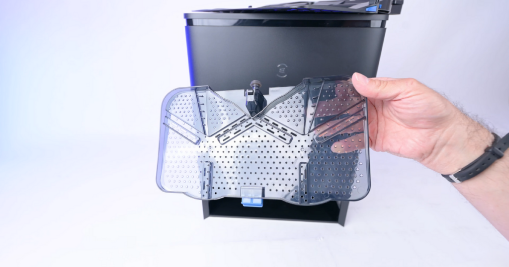 A hand holding the removable tray from the Ecovacs Deebot T30S Omni Station, highlighting its design and perforations, with the Omni Station in the background.