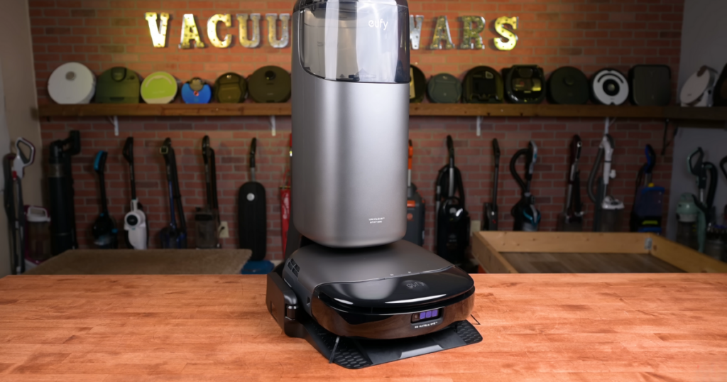 Eufy S1 Pro Omni robot vacuum and UniClean Station displayed on a wooden table in front of a brick wall with various vacuum models hanging in the background and a "Vacuum Wars" sign above.