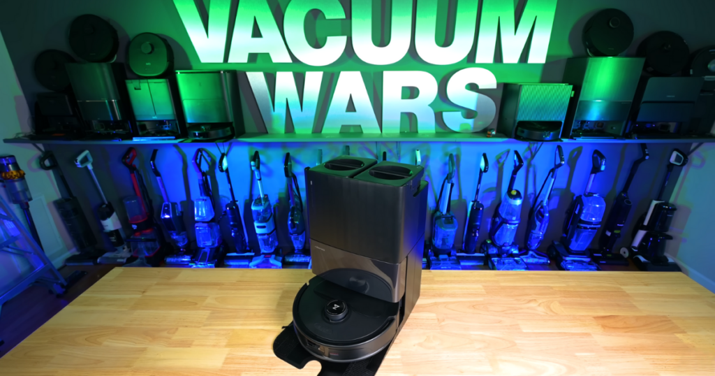 Roborock Qrevo Master robot vacuum and mop displayed on a wooden surface with various vacuum cleaners in the background and the "Vacuum Wars" sign illuminated above.