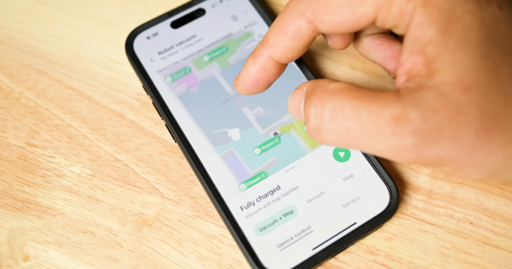 A close-up of a hand interacting with a smartphone displaying the map interface of the Samsung Bespoke Jet Bot Combo AI robot vacuum app, showing different rooms and cleaning options.