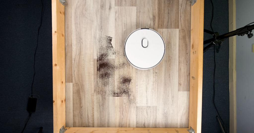 A top-down view of a Samsung robot vacuum cleaning a wooden floor inside a wooden-framed area, picking up a concentrated pile of dirt or debris.