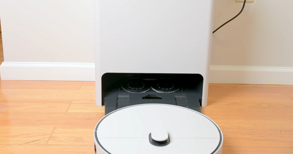 The Samsung Bespoke Jet Bot AI Combo robot vacuum approaching its all-in-one cleaning station on a hardwood floor, with the mop pads visible inside the station.