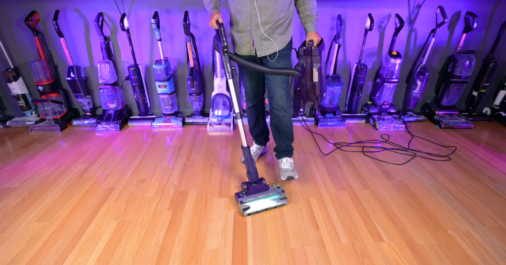 A person using the Shark PowerDetect vacuum in Powered Lift-Away mode on a hardwood floor. The vacuum base is detached and being maneuvered with the wand, while the canister is held in the other hand. A row of various vacuum models is visible in the background.