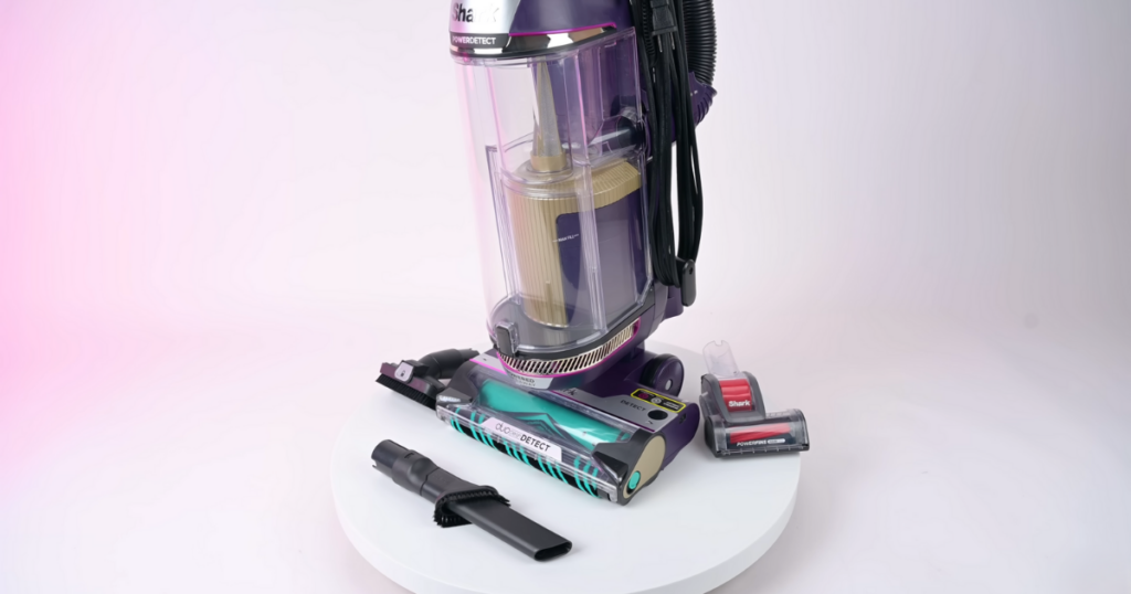 The Shark PowerDetect vacuum cleaner is displayed along with its various attachments, including a crevice tool, brush attachment, and motorized brush. The vacuum and accessories are arranged neatly on a white platform against a light pink background.