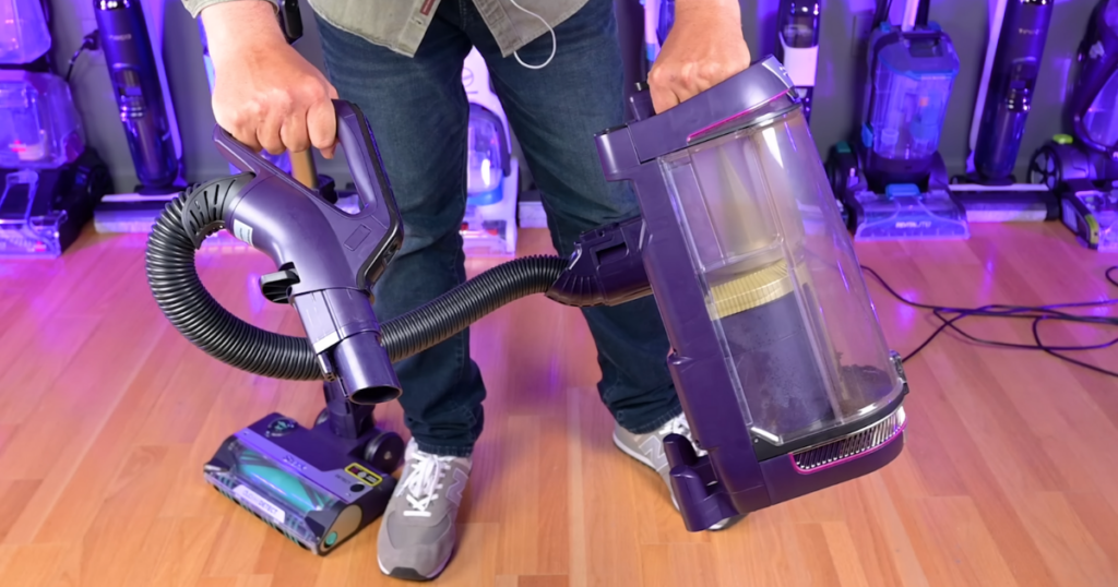 Person demonstrating the Shark PowerDetect vacuum's Lift-Away feature, holding the detached canister in one hand and the hose handle in the other. The vacuum base remains on the floor.