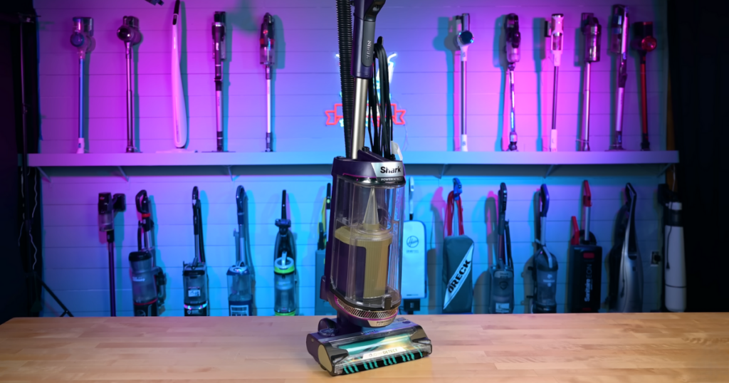 The Shark PowerDetect vacuum cleaner is displayed prominently on a wooden surface, with a variety of other vacuum models mounted on the wall in the background. The room is illuminated with blue and purple lighting.