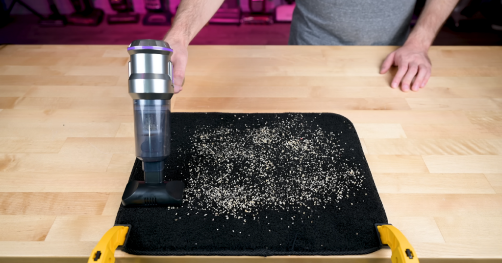 A person uses the PeroBuno mini vacuum cleaner on a black mat covered with debris, demonstrating the vacuum's cleaning performance during an evaluation.