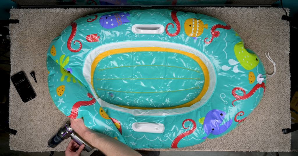 A colorful inflatable raft featuring cartoon sea creatures is being inflated with a PeroBuno 2-in-1 vacuum cleaner. A smartphone with a timer showing 31.44 seconds is placed nearby on the carpeted surface.