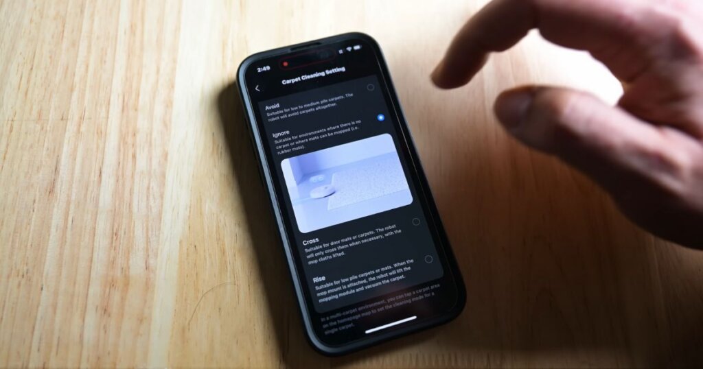 A close-up of a smartphone displaying the Roborock Qrevo Edge app, highlighting the carpet cleaning settings with options like Avoid, Ignore, Cross, and Rise while a hand hovers near the screen.
