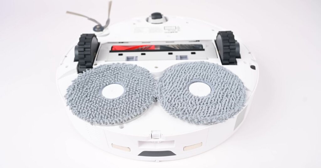 A close-up view of the underside of the Roborock Qrevo Edge showing its dual spinning mop pads, side brush, and DuoDivide brush roll. The mop pads are circular and positioned at the back of the vacuum.