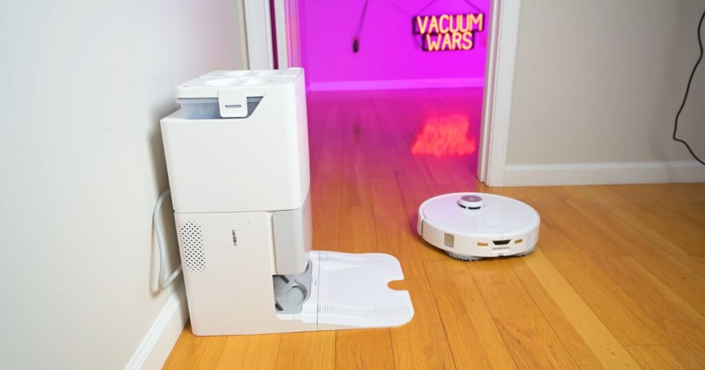 Roborock Qrevo Edge robot vacuum leaves its dock station to clean, with a "Vacuum Wars" neon sign glowing in the background.