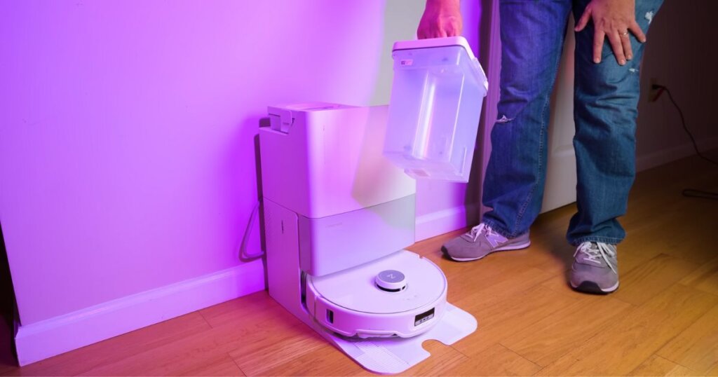 A person removing one of two water tanks from the Roborock QRevo Edge docking station. The docking station is placed against a wall, and the scene is lit with purple ambient lighting.