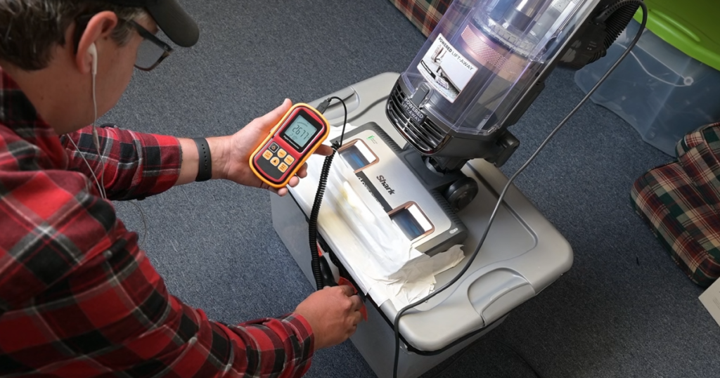 A person measuring airflow at the floorhead of a Shark Vertex upright vacuum using a digital anemometer.