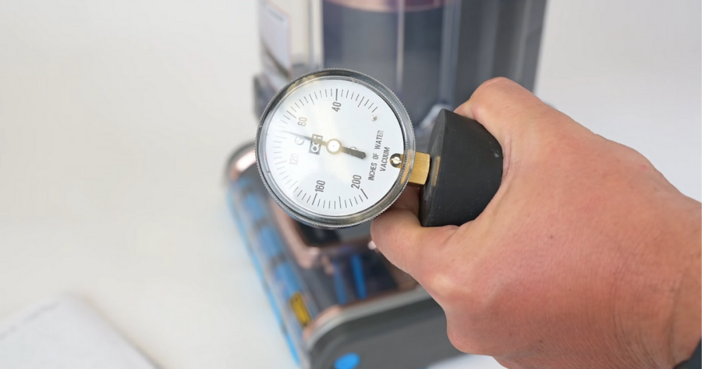 Close-up of a hand holding a suction gauge at the hose of a Shark Vertex upright vacuum to measure water lift in inches.