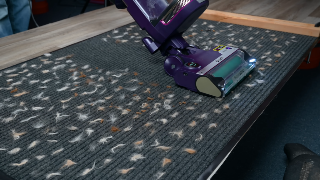 Upright vacuum cleaner on a test mat covered with pet hair and debris, demonstrating hair pickup performance.