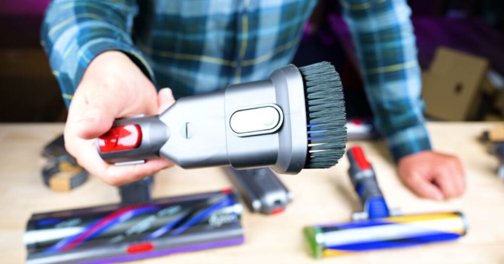 Hand holding a Dyson-style dusting brush with long bristles, surrounded by various other vacuum attachments.