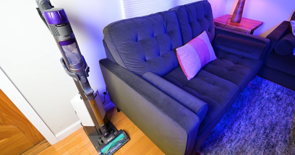 Cordless vacuum with an auto-emptying bin system, shown docked next to a sofa in a living room.