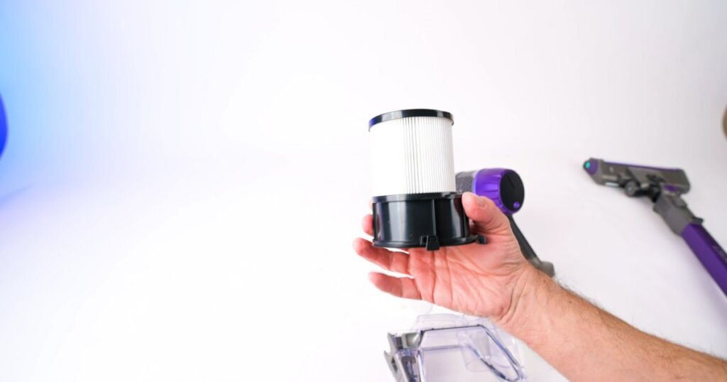 Close-up of a sealed HEPA filter being held in front of a cordless vacuum with the dustbin detached.