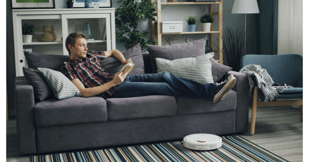 A man quietly reading while a robot vacuum is rolling by.