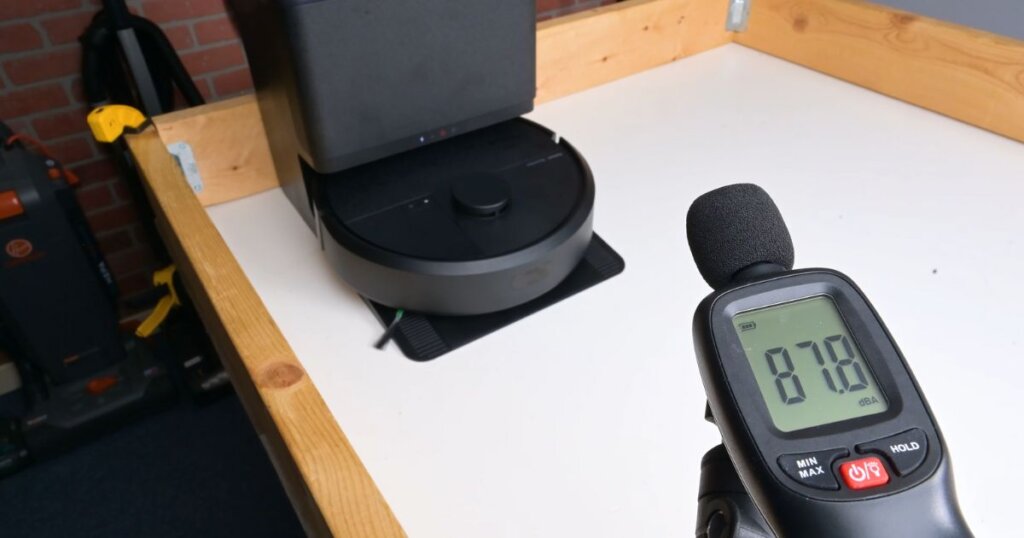 Roomba Plus 405 Combo dock being tested for noise output during emptying, showing 87.8 dBA on a sound meter.