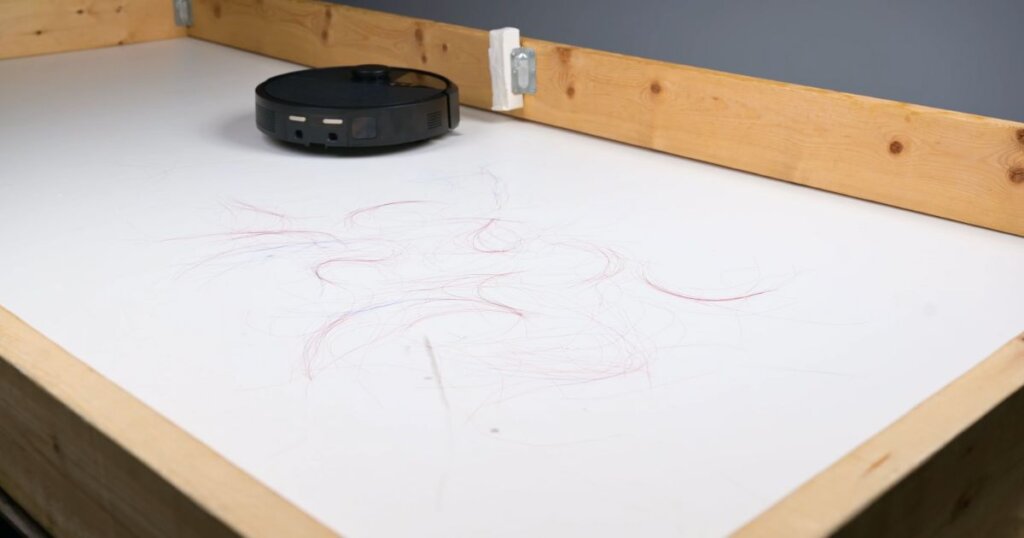 Robot vacuum on a white test surface with strands of red and dark hair scattered, mid-hair pickup test.