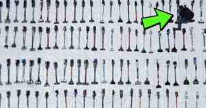 Close to 100 cordless stick vacuums are arranged in rows on a white tarp, with a person kneeling among them and a large green arrow pointing to one vacuum.