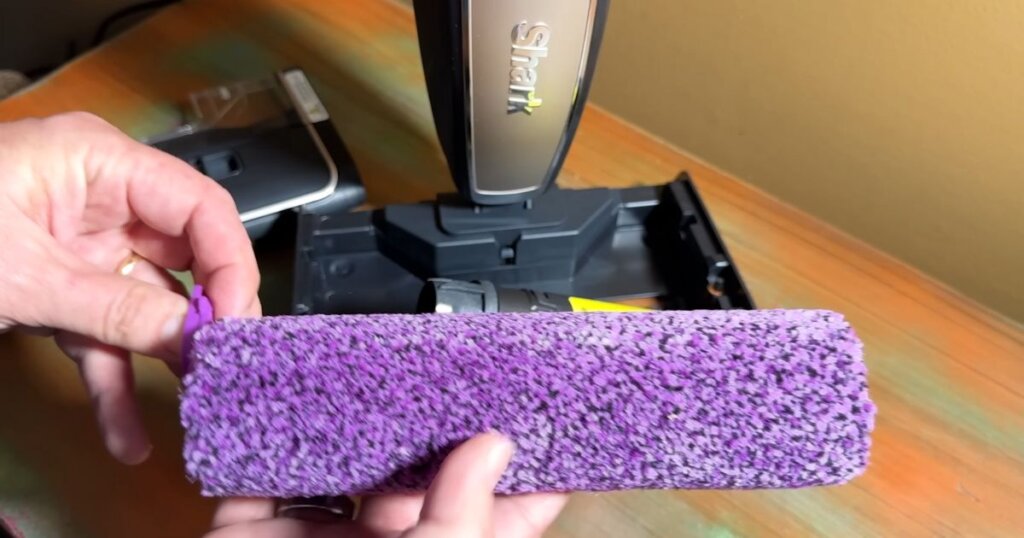 Close-up of a hand holding the purple roller brush of the Shark Steam Pickup in front of the vacuum's base unit.