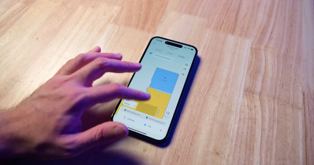 A hand operating the MOVA app on a smartphone, showing mapped cleaning zones with yellow and blue overlays.