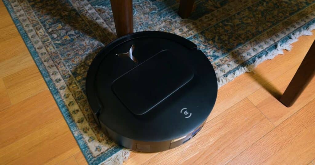 Ecovacs T80 Omni robot vacuum caught under a wooden table leg during cleaning on a patterned rug over hardwood flooring.