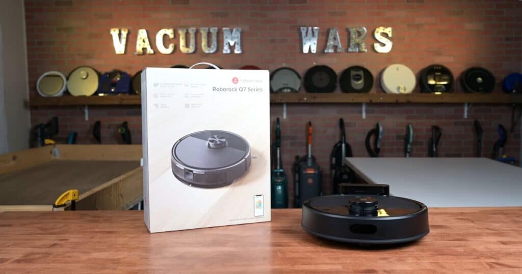 Roborock Q7 M5 robot vacuum and retail box on a workbench in the Vacuum Wars studio with various vacuums on a wall rack in the background.