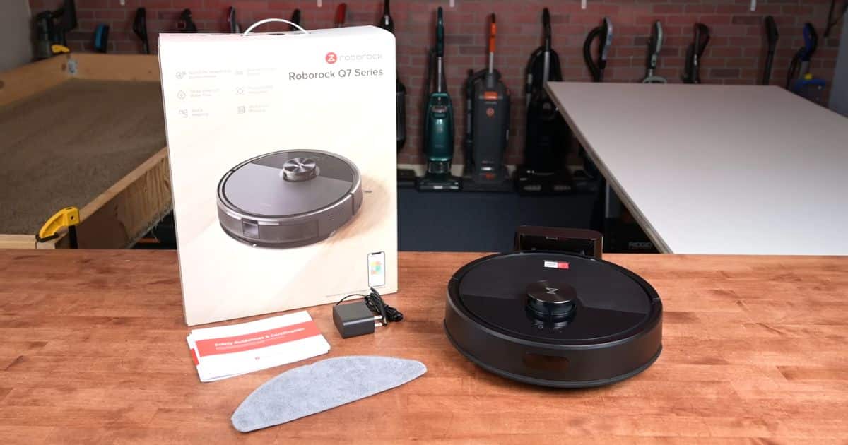 Roborock Q7 M5 robot vacuum, box, charger, mop pad, and manuals arranged on a workbench in the Vacuum Wars studio.