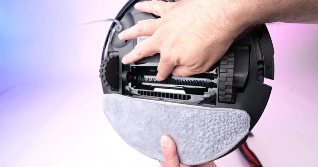 Underside of the Roborock Q7 M5+ with mop pad attached; hand points at the roller area showing the jaw-style comb above the main brush.