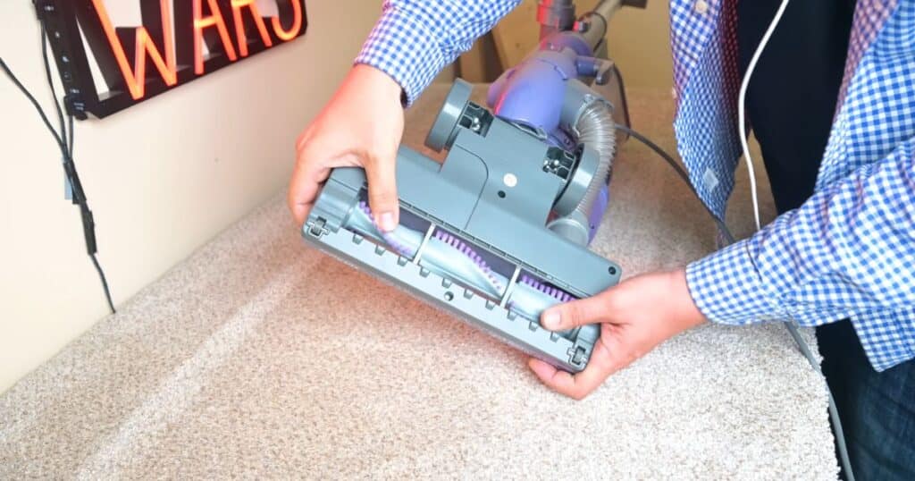 Hands holding the underside of a vacuum floorhead showing brushroll design on a carpeted floor.