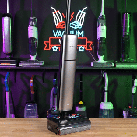 Dreame Aero Pro in studio 450 ©Vacuum Wars | Vacuum Wars Cordless stick vacuum standing on its charging dock on a wooden table, with a neon-lit showroom backdrop.
