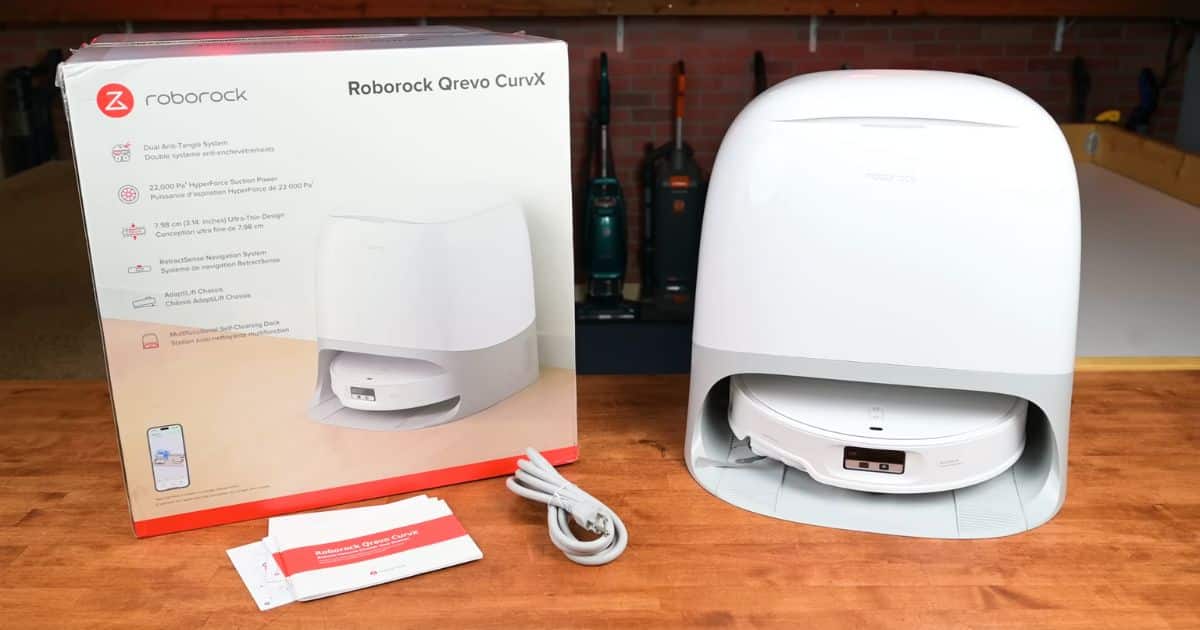 Roborock Qrevo CurvX robot vacuum and its packaging shown on a wooden table, including power cord and manuals.