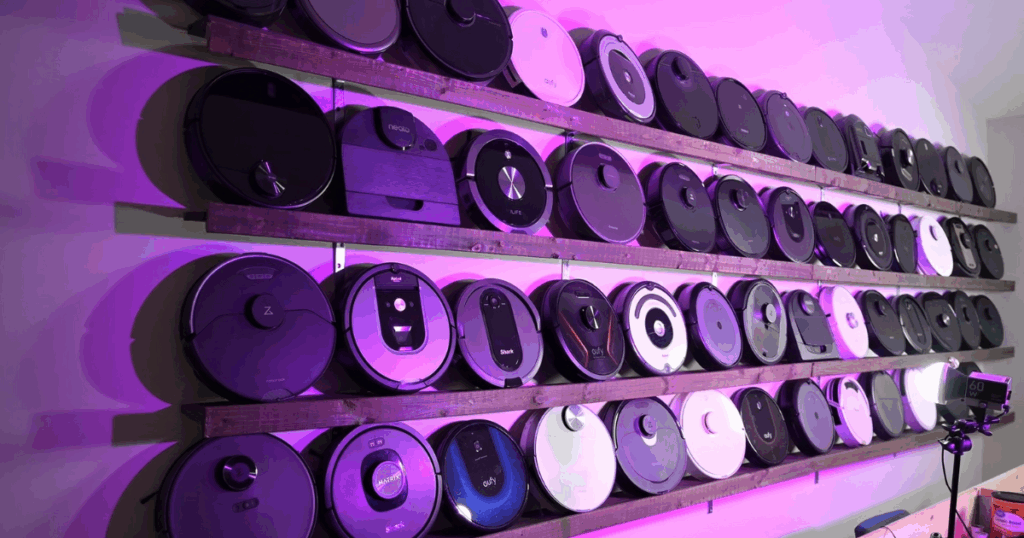 Robot Vacuums at Vacuum Wars | Vacuum Wars A wall display at Vacuum Wars showcasing dozens of robot vacuums from various brands, neatly arranged on wooden shelves and illuminated by purple lighting.