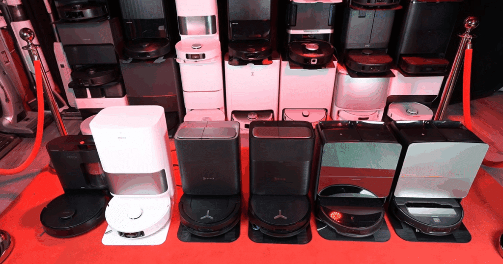 A lineup of robot vacuums with their auto-empty and multi-functional docks displayed on a red carpet, featuring models from leading brands like Roborock, Ecovacs, Dreame, and Yeedi.