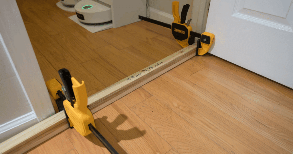 Threshold climbing test | Vacuum Wars A close-up of a robot vacuum threshold climbing test setup, featuring a 44mm (1.75-inch) wooden barrier secured with clamps on a hardwood floor. This setup is used to measure how high robot vacuums can climb over obstacles during Vacuum Wars’ standardized performance testing.