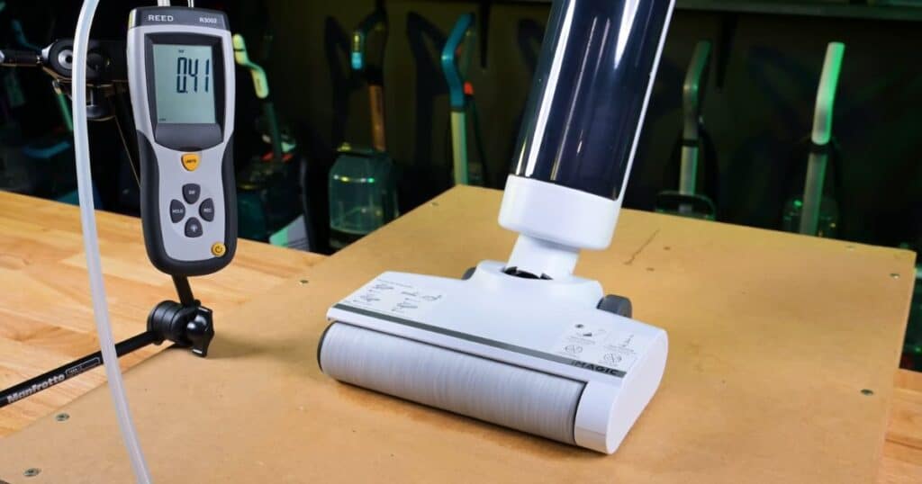 A suction test setup showing a hard floor cleanerโs roller head placed under a meter connected to tubing, used to measure usable suction at the floorhead.