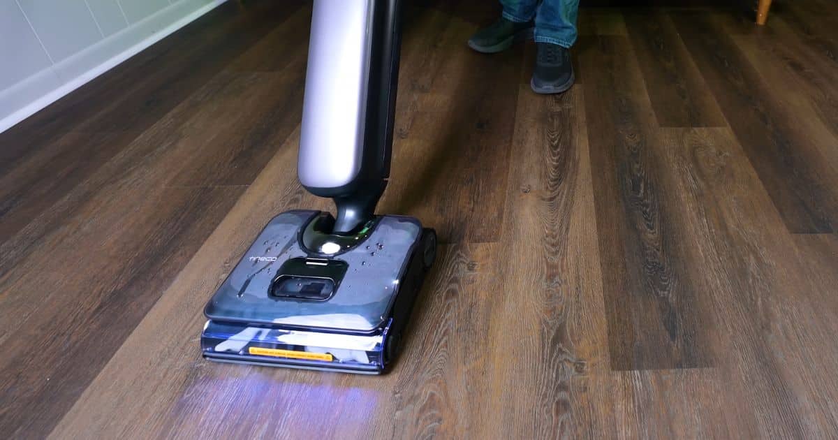 A hard floor cleaner with visible steam rising from the floorhead as it moves across a dark wood floor during operation.