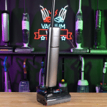 Dreame Aero Pro in studio 150 ©Vacuum Wars | Vacuum Wars Cordless stick vacuum with a transparent dustbin and long handle, standing upright on a wooden counter; colorful bottles on a backlit display shelf behind.