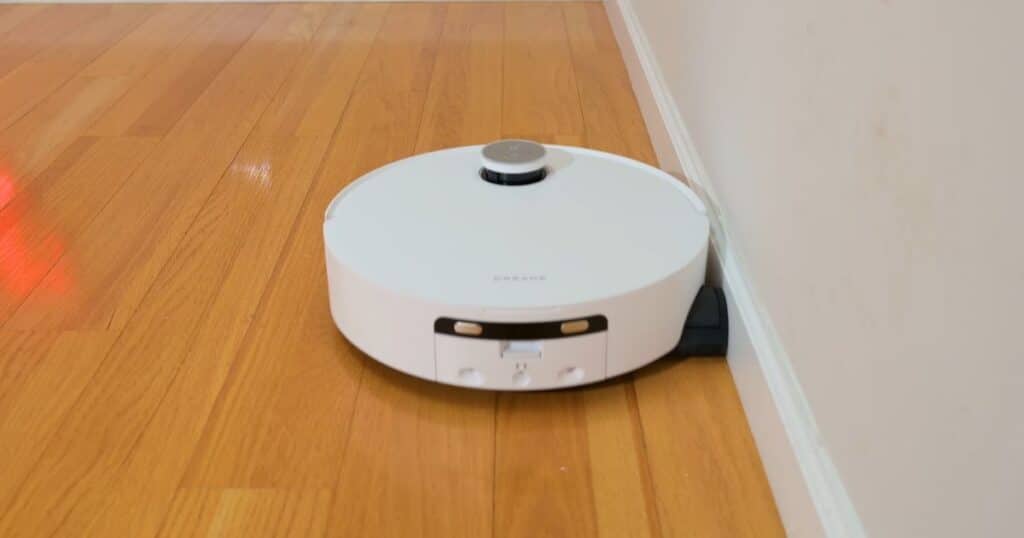 | Vacuum Wars The Dreame Aqua 10 Ultra Roller robot vacuum cleaning along a wall, demonstrating its edge-extending roller mop design that automatically extends to reach baseboards and corners for more thorough cleaning coverage.