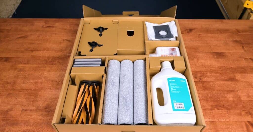 Box contents of the MOVA Z60 Ultra Roller Complete robot vacuum showing multiple replacement accessories including three roller mops, two side brushes, extra main brushes, filters, a bottle of cleaning solution, and dust bags arranged neatly in cardboard packaging.