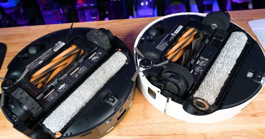 Bottom view comparison of two robot vacuums placed side by side on a wooden table — the black MOVA Z60 Ultra Roller on the left and the white Dreame Aqua10 on the right. Both show dual-roller brush assemblies, side brushes, and roller mop attachments.